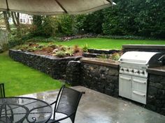 an outdoor kitchen and grill area with grass in the back ground, next to a stone wall