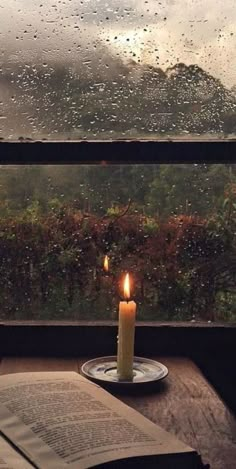 a lit candle on a plate next to an open book and window with raindrops