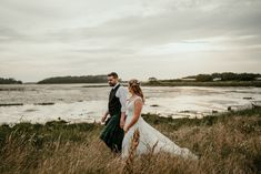 a bride and groom walking through tall grass by the water