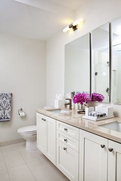 a white bathroom with two sinks, mirrors and flowers in vases on the counter