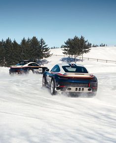 two sports cars driving through the snow in front of some pine trees on a sunny day