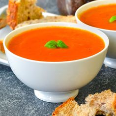 two bowls of tomato soup with bread on the side and basil garnishes