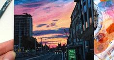 someone is holding up a painting in front of a cityscape at sunset with buildings and street lights