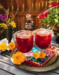 two glasses filled with red liquid sitting on top of a wooden table next to flowers