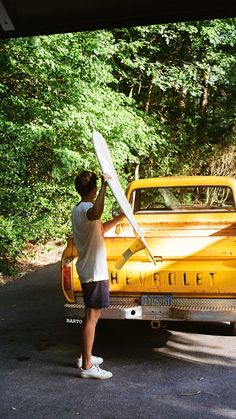 a man holding a white frisbee standing next to a yellow truck with trees in the background