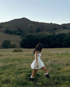 a woman in a white dress walking across a lush green field with mountains in the background