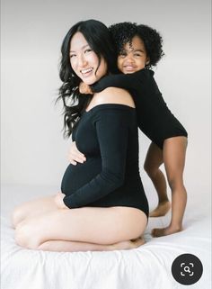 two women in black bodysuits sitting on a white bed and posing for the camera