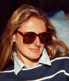 a woman wearing sunglasses and a sweater looks off to the side with her hair blowing in the wind