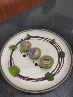 a white plate topped with three desserts covered in purple and green topping on top of a table