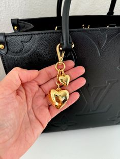 a hand holding a black purse with two gold hearts hanging from it's handles