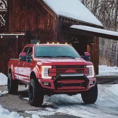 a red truck is parked in front of a barn with snow on the ground and trees