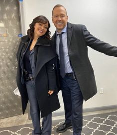 a man and woman standing next to each other in front of a white wall wearing coats