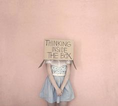 a woman standing in front of a pink wall with a sign on her head that says thinking inside the box