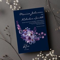 a wedding card with purple flowers on it next to some white flowers and branches in the background