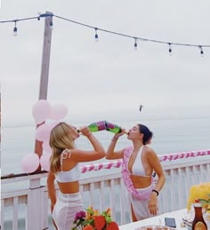 two women in bathing suits drinking from bottles on a balcony overlooking the water and balloons