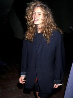 a woman with long curly hair wearing a black blazer and red skirt is smiling at the camera