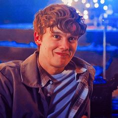 a young man with curly hair wearing a jacket and striped shirt smiling at the camera