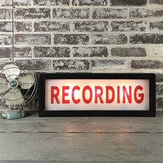 a sign that says recording on it next to a fan and a brick wall in the background