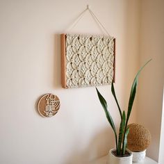 two plants are sitting on a table next to a wall hanging with a woven piece of art