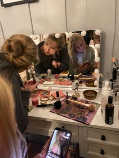 three girls looking at their cell phones in front of a vanity with makeup on it