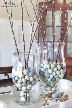 two vases filled with eggs and twigs on top of a white tablecloth covered tray