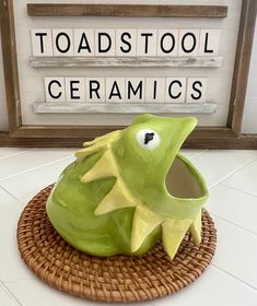 a green ceramic fish sitting on top of a wicker basket next to a sign that reads toad's tool ceramics