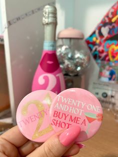 someone holding two pink and gold buttons in front of a bottle with the words twenty - one and hot buy me a shot on it