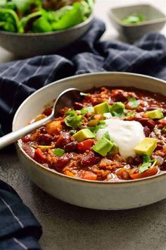 a bowl of chili with sour cream and avocado on top, next to a bowl of lettuce