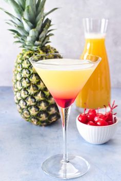 two glasses filled with different colored drinks next to pineapples and cherries on a table