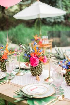 an outdoor table set with pineapples, flowers and plates for a tropical themed party