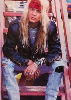 a man with long hair sitting on top of a wooden ladder wearing jeans and a leather jacket