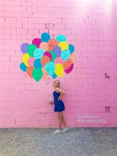 a woman standing in front of a pink brick wall holding a bunch of colorful balloons