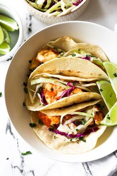 three fish tacos in a white bowl with lime wedges on the side and another dish next to it