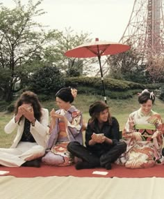 four women sitting on the ground in front of an umbrella and one woman is holding her hand up to her face