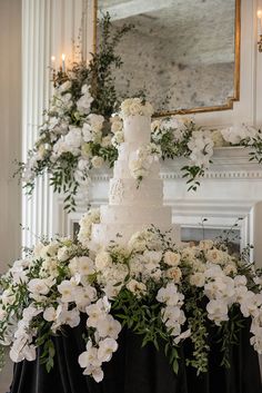 a wedding cake with white flowers and greenery in front of a fireplace mantel