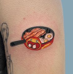 a woman's arm with a tattoo on it that has an image of bacon and eggs in a frying pan