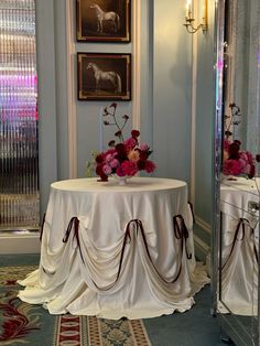 a table with flowers on it in front of a mirror and two framed pictures behind it