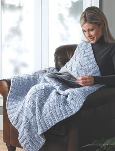 a woman sitting in a chair holding a book and wearing a knitted blanket over her lap