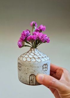 a hand holding a small white vase with purple flowers in it's top half