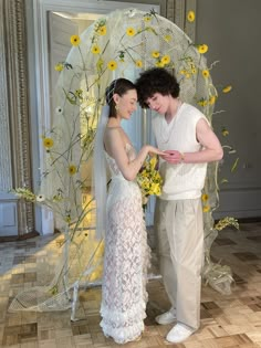 a man and woman standing next to each other in front of a white umbrella with yellow flowers on it