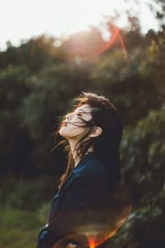 a woman with her eyes closed standing in front of trees and sun flares behind her