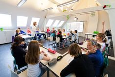 a group of children sitting at desks in a room with white walls and windows