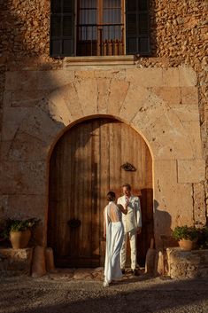 a man and woman standing in front of a stone building with an open door that has shutters on it