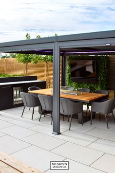 Elevate your outdoor gatherings with this sleek, contemporary dining setup. Featuring a pergola with integrated lighting, a built-in kitchen, and a lush green wall with a TV for entertainment, this space is perfect for hosting in style. Designed for both comfort and functionality by The Garden Design Co. Transform your garden today! #gardendesign #moderngarden #outdoorkitchen #gardenideas