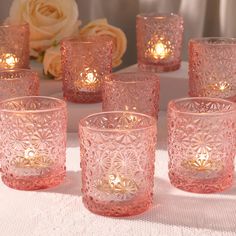 many pink glass cups with lit candles on a white table cloth and flowers in the background