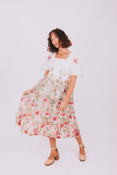 a woman wearing a white dress with pink flowers on it and brown shoes, standing in front of a white background