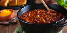 a pot filled with chili and bread on top of a wooden table next to other foods