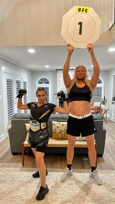 two women standing in a living room with one holding up the number 1 on her chest