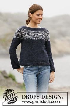 a woman standing in front of a body of water wearing a black sweater and jeans