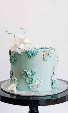 a blue cake with white flowers on top sitting on a black stand in front of a white wall